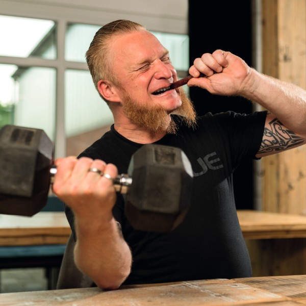 Mann mit Bart und Hanteln beißt in eine Mini-Salami